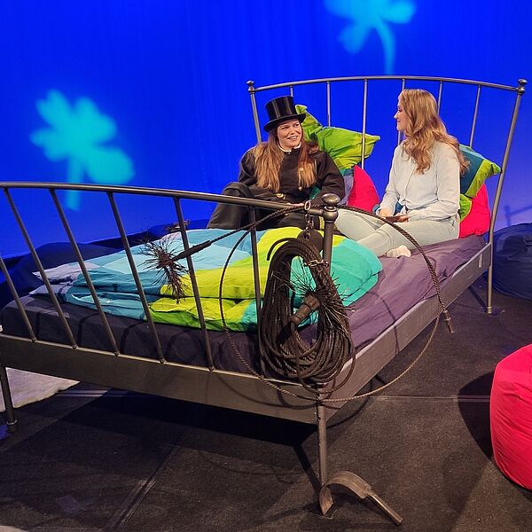 Moderatorin und eine Schornsteinfegerin, die zu Gast in der Sendung ist, sitzen auf einem Bett