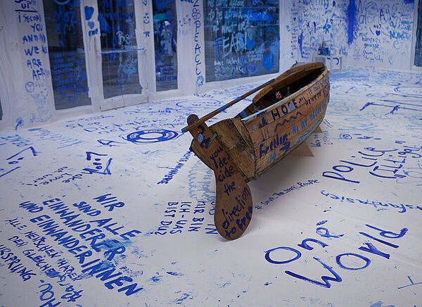 Die ersten Besucher haben bereits in blauer Farbe ihre Gedanken zum Thema Migration auf dem Holzboot und an den weißen Wänden hinterlassen