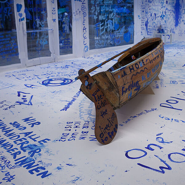 Die ersten Besucher haben bereits in blauer Farbe ihre Gedanken zum Thema Migration auf dem Holzboot und an den weißen Wänden hinterlassen