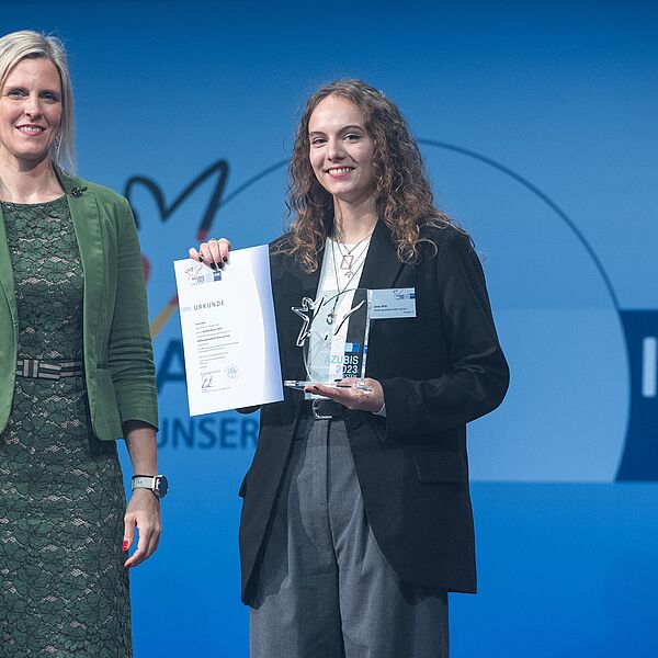 Swaantje Creusen, Vorsitzende des DIHK-Bildungsausschusses, und Jana Witt stehen nebeneinander auf der Bühne. Jana Witt hält ihre Urkunde hoch. 