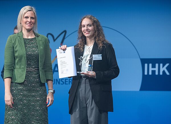 Swaantje Creusen, Vorsitzende des DIHK-Bildungsausschusses, und Jana Witt stehen nebeneinander auf der Bühne. Jana Witt hält ihre Urkunde hoch. 