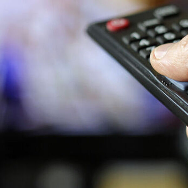 Hand, die eine Fernbedienung hält. Im Hintergrund läuft der Fernseher.