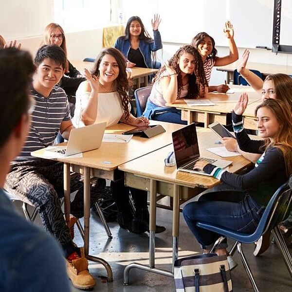 Klassenzimmer, in dem Schüler*innen an Tischen sitzen, von denen einige sich per Handzeichen melden